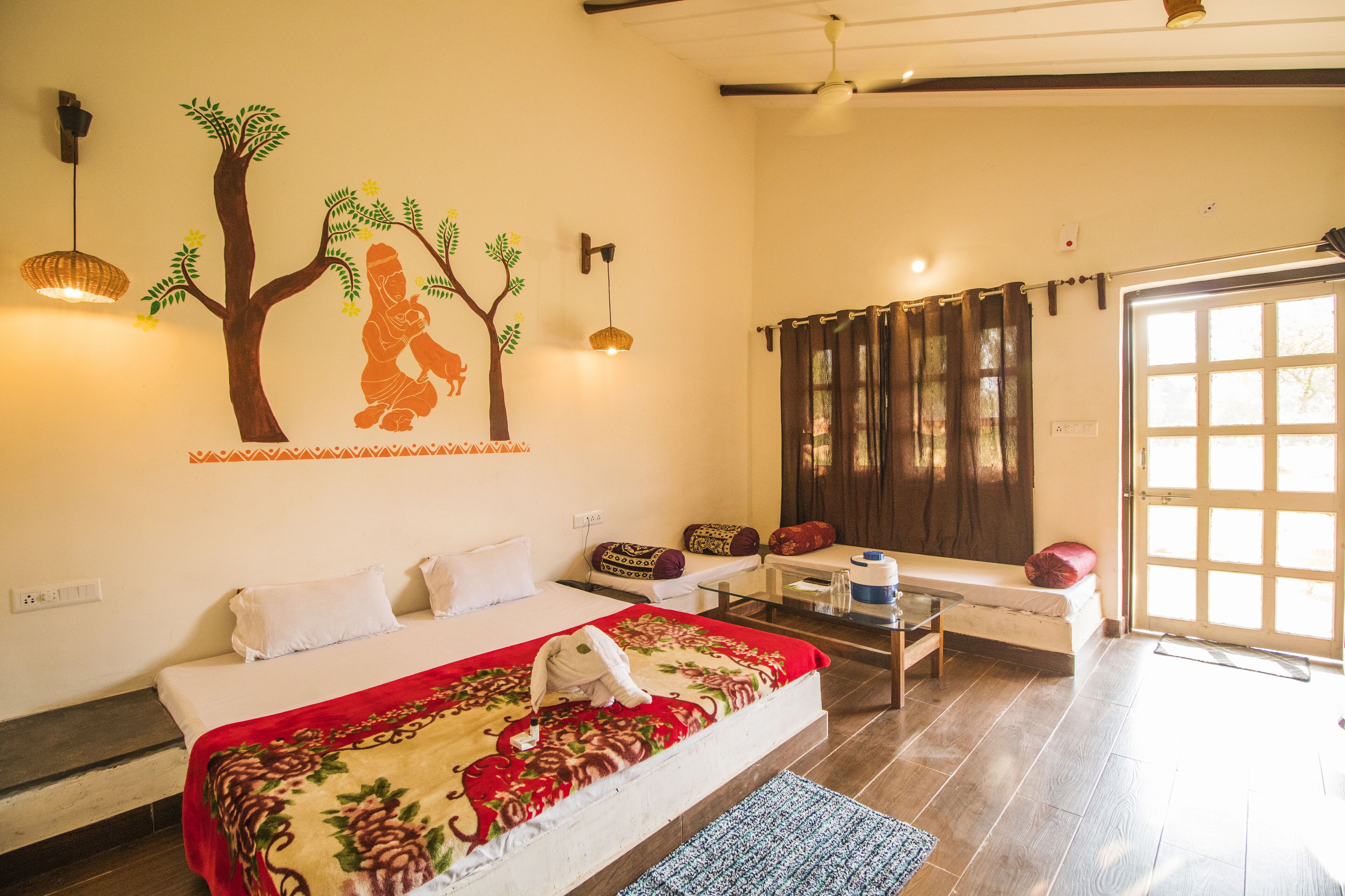 A bedroom with a bed, a small table, a sofa, a wall mural, a window, and a door.