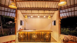A bar area with a wooden counter, hanging wicker lamps, and a view of the outside. - Pachmarhi Foothill Cottages by Vanyum (Pipariya)
