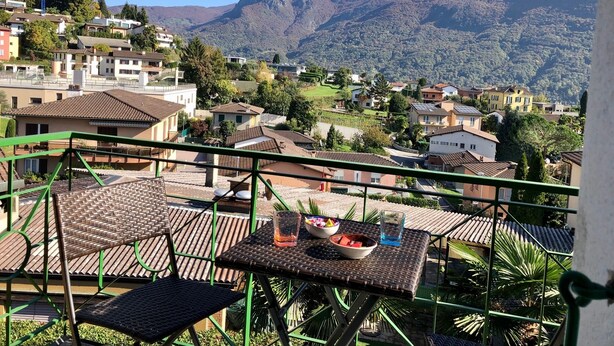 A balcony with wicker furniture, a table with a bowl of fruit and a glass, and a view of a hillside village with houses and mountains. - Top Ferienwohnung - Ticino Lugano (Comano) (Comano)