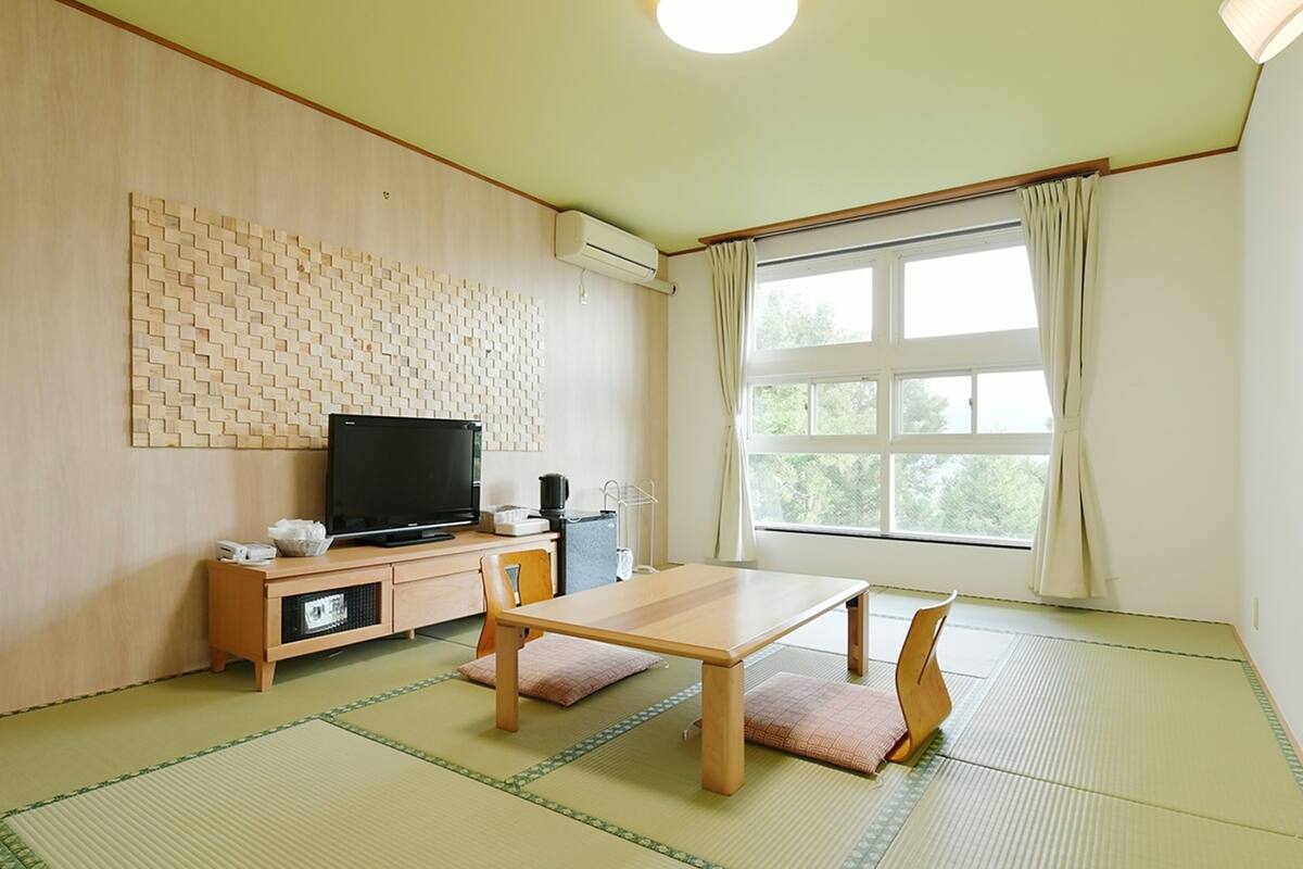 A room with tatami mat flooring, a wooden table, a television on a cabinet, and a window with curtains.