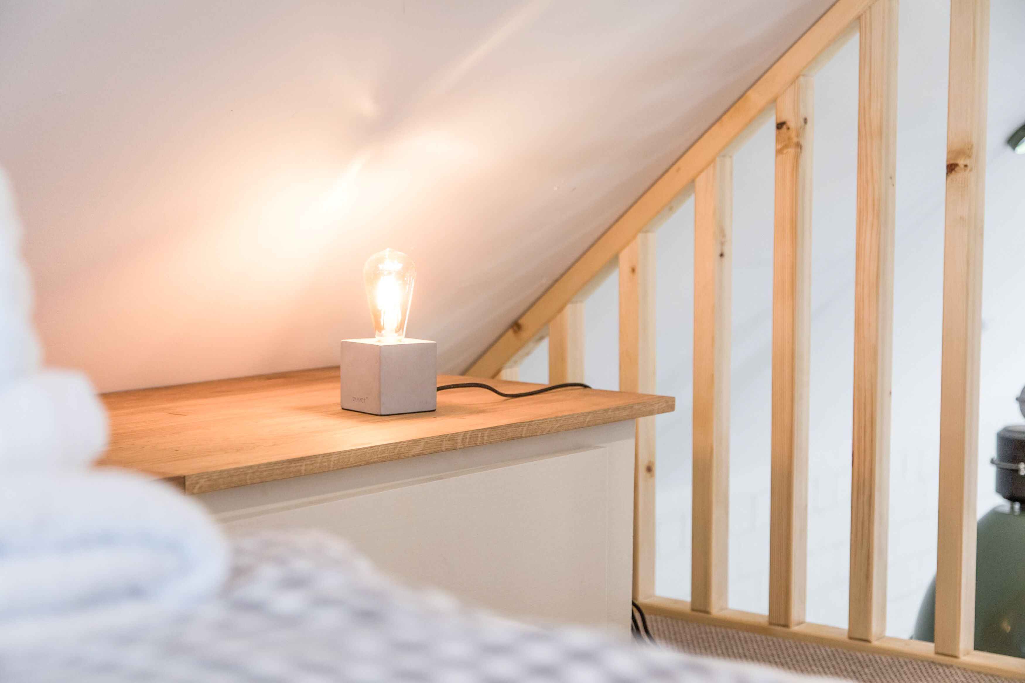 Une pièce avec un plafond en pente, un escalier en bois et une petite table sur laquelle se trouve un luminaire.
