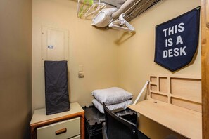 Une petite pièce comprenant un bureau, une chaise, un lit avec des oreillers, un porte-manteau mural et une pancarte indiquant : « ICI SE TROUVE UN BUREAU ».