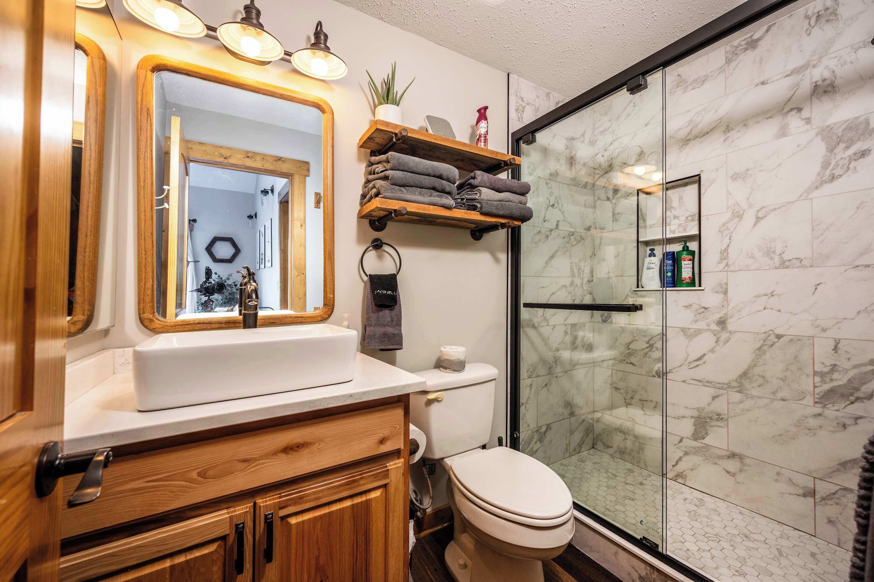 Un baño con tocador de madera, un lavamanos, inodoro, ducha con puertas de vidrio y repisa con toallas.