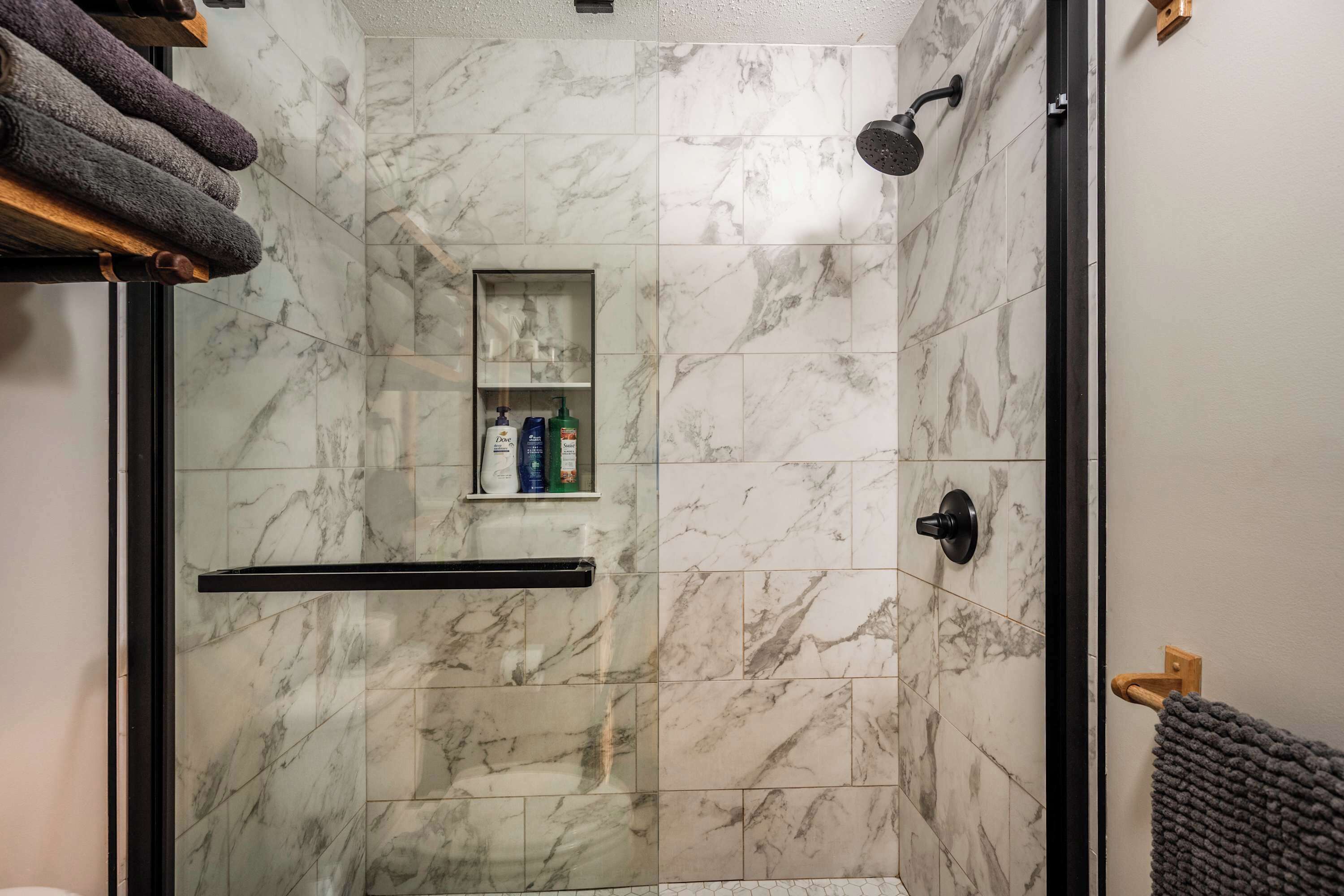 A shower with a glass door, marble tiles, a shelf with toiletries, and a towel rack.