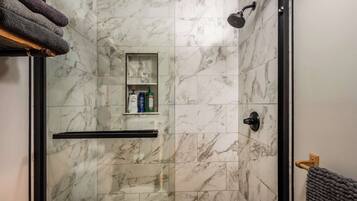 A shower with a glass door, marble tiles, a shelf with toiletries, and a towel rack.