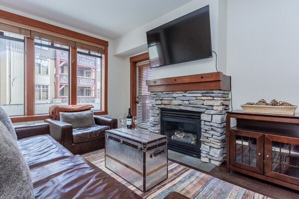 A living room with a stone fireplace, a television mounted above it, and a large window with blinds.