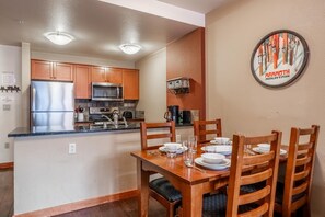 A compact kitchen with wooden cabinets, a stainless steel refrigerator, and a microwave. There is a dining table set with plates, glasses, and cutlery. A coffee maker and kettle are on the counter.