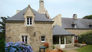 A stone house with a traditional design, featuring a stone chimney, a window with a white frame, and a wooden table and chairs in the front yard. - The Closerie du Manoir for 7 people (Audierne)