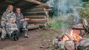 Two people sitting outside a log cabin with a campfire.