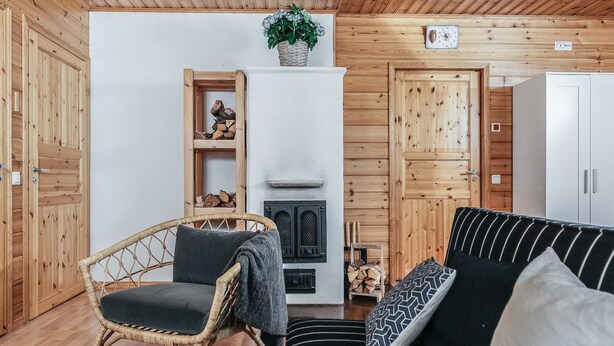 A cozy room with a fireplace, wooden walls, a wicker chair, a striped sofa, and a small shelf with logs. - Vacation home B by Interhome (Porvoo)