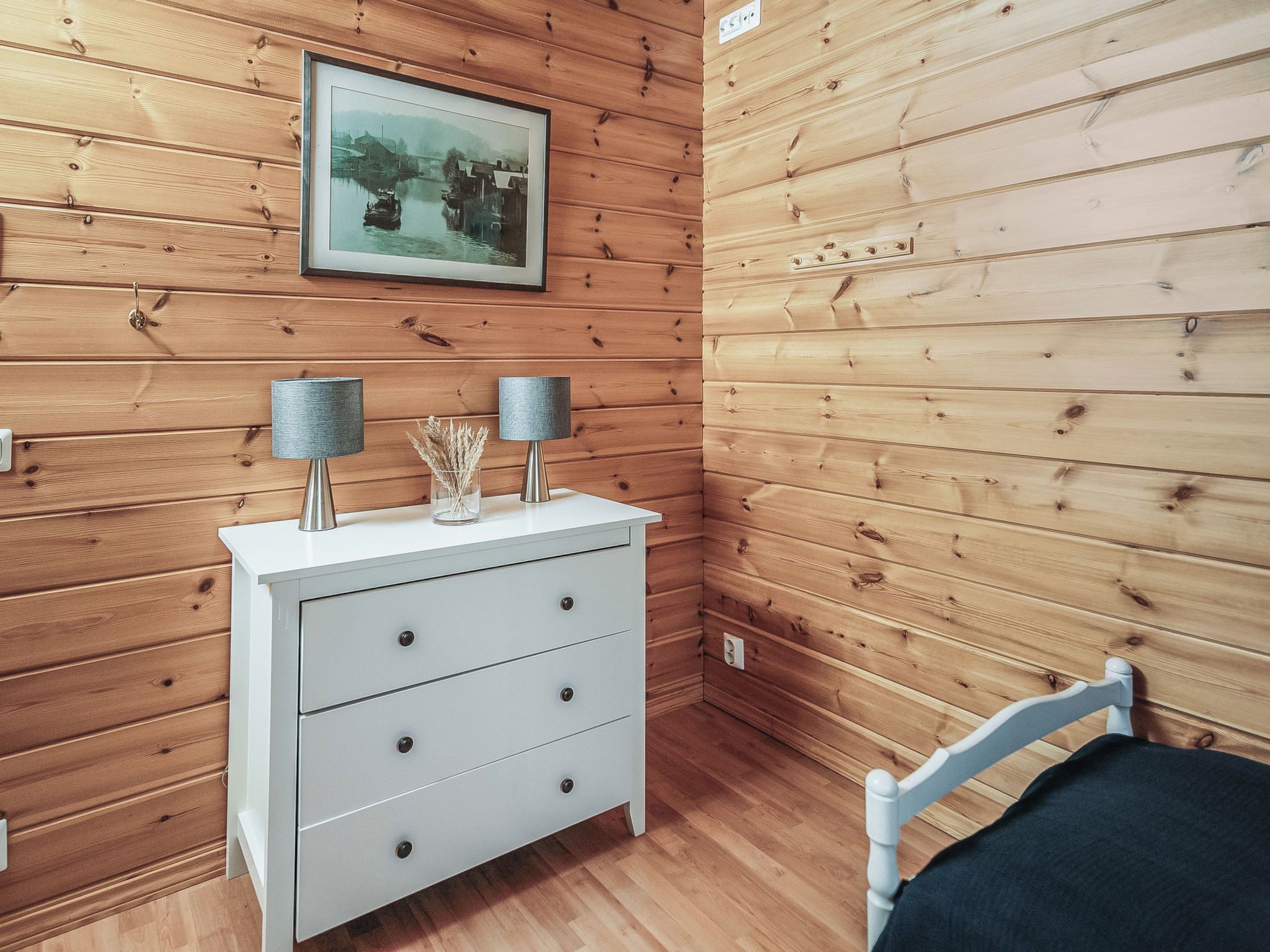 A room with wooden panel walls, a white dresser with drawers, two lamps, a framed picture on the wall, and a white chair.