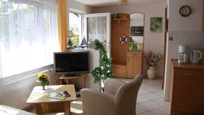 Ett vardagsrum med en tv, en soffa, ett matbord med böcker, en köksdel med en kaffebryggare och ett fönster med gardiner.