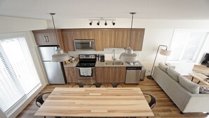 Una cocina moderna con armarios de madera, electrodomésticos de acero inoxidable, una mesa de comedor con sillas y un sofá en una habitación luminosa con ventanas amplias.