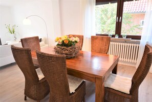 Ein Essbereich mit Holztisch, Korbstühlen, einem gewebten Korb mit Blumen, einer Stehlampe und Blick auf ein Backsteingebäude durch ein Fenster.