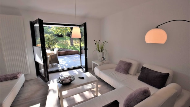 A modern living room with a white sofa, a coffee table, and a view of a garden through an open door. - Vacation apartment T & T 3 (Malente)