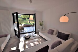 Ein modernes Wohnzimmer mit einem weißen Sofa, einem Couchtisch und Blick auf einen Garten durch eine offene Tür.