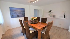 A dining area with a wooden table and wicker chairs, a wall-mounted painting, and a shelf with decorative items. - Vacation apartment T & T 3 (Malente)