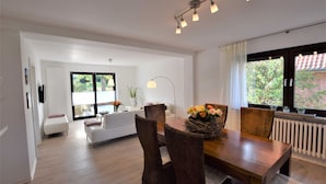 A modern living room with a dining area, featuring a wooden table, wicker chairs, and a large window with a view of greenery. - Vacation apartment T & T 3 (Malente)