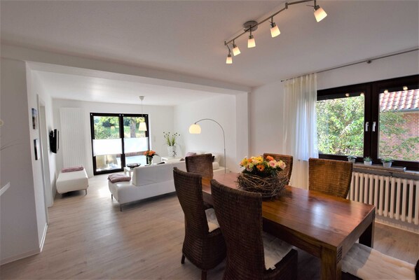 Ein modernes Wohnzimmer mit Essbereich, ausgestattet mit einem Holztisch, Korbstühlen und einem großen Fenster mit Blick auf Grünflächen.