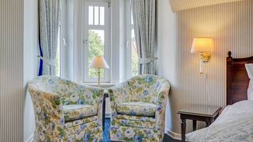 A hotel room with two floral armchairs, a bed, a nightstand, a lamp, and a window with curtains.