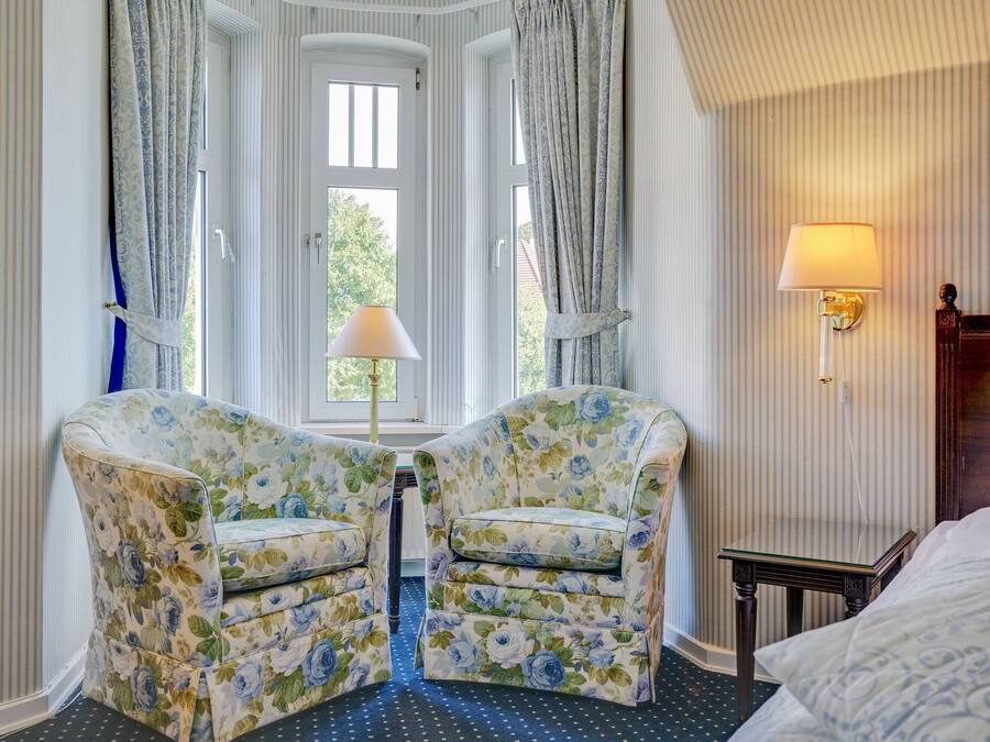 A hotel room with two floral armchairs, a bed, a nightstand, a lamp, and a window with curtains.
