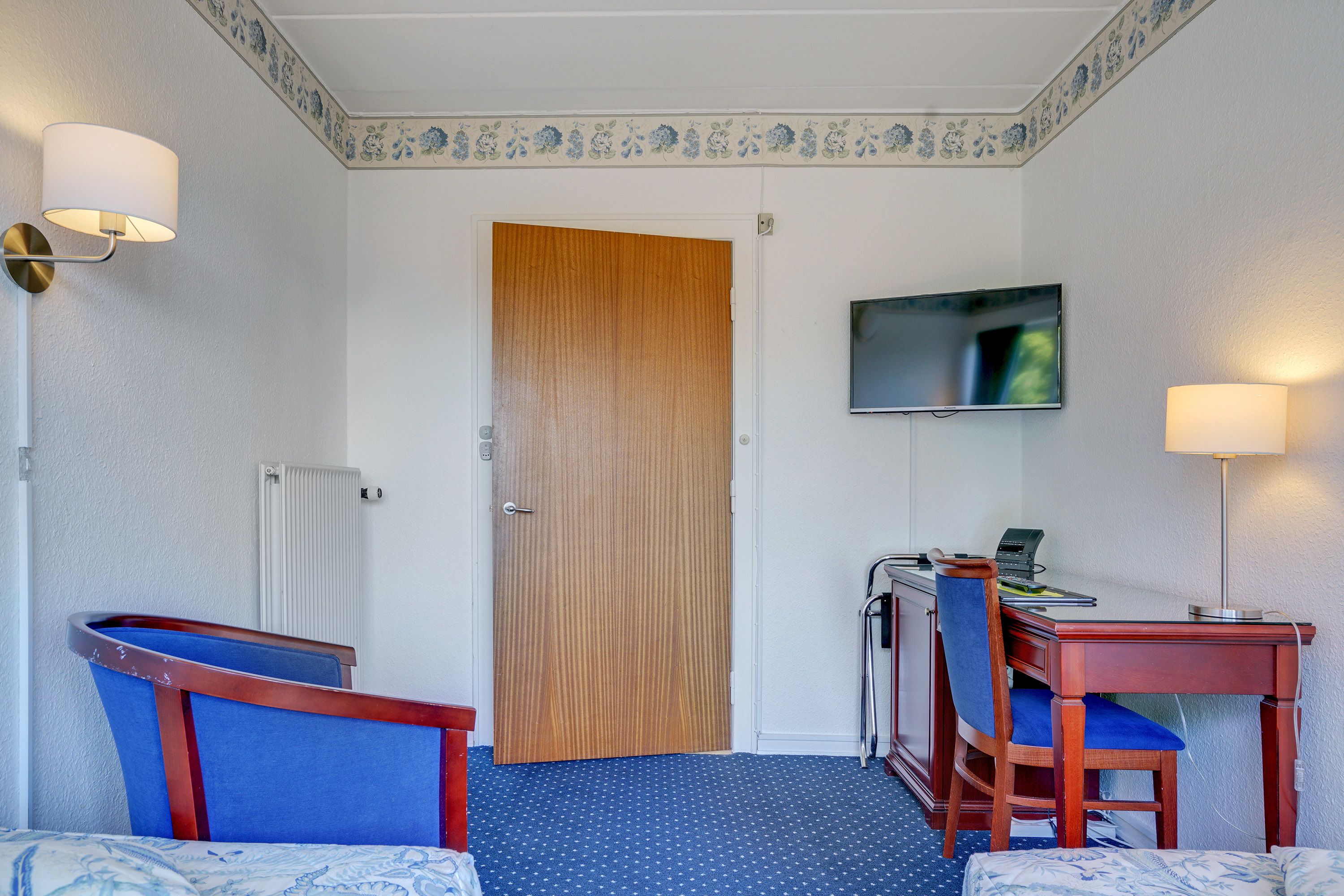 Une chambre d’hôtel avec une porte en bois, un bureau avec un ordinateur, deux chaises, un téléviseur fixé au mur et une moquette à motifs.