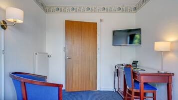 Une chambre d’hôtel avec une porte en bois, un bureau avec un ordinateur, deux chaises, un téléviseur fixé au mur et une moquette à motifs.