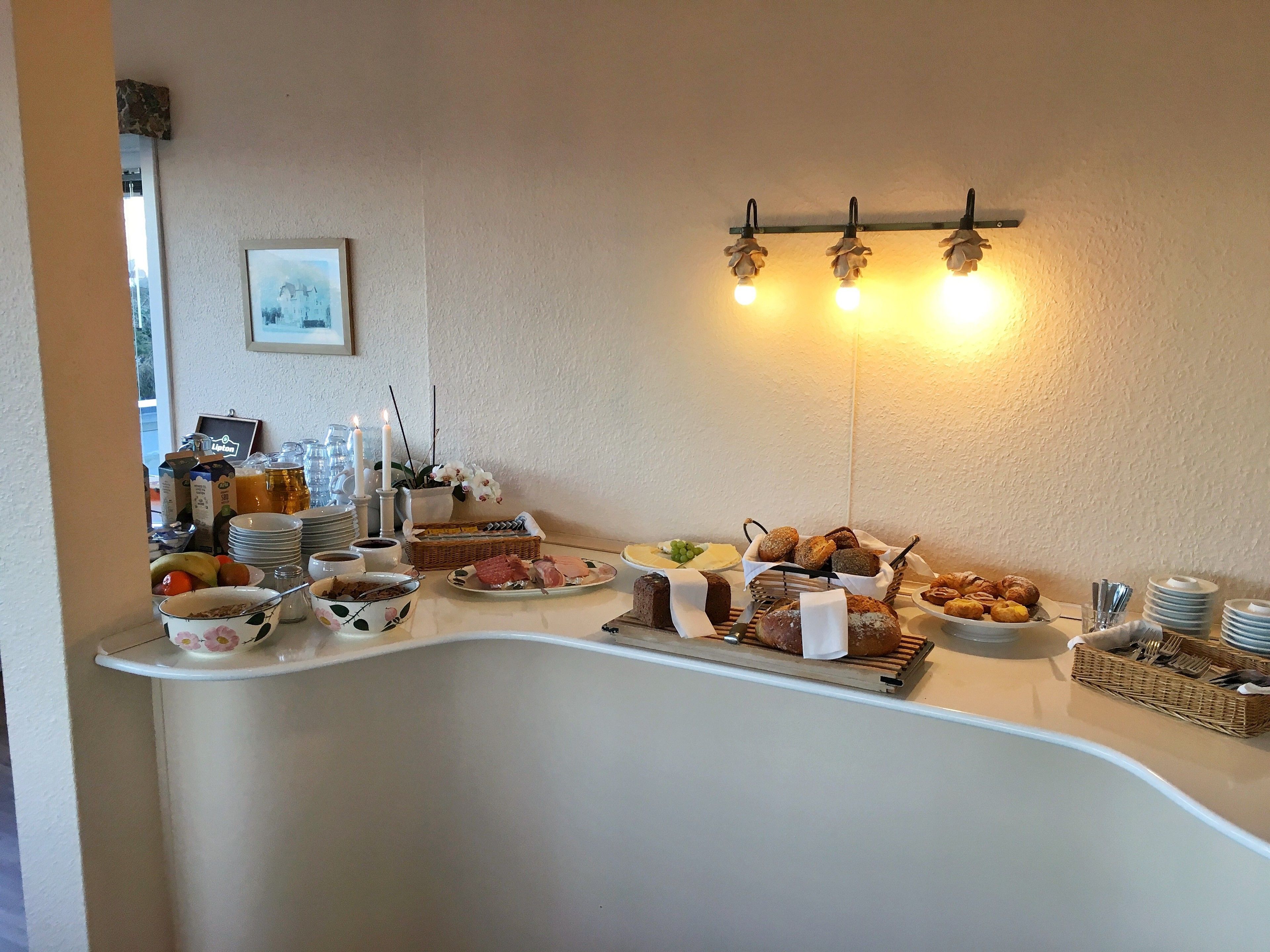 Una mesa de buffet con diversos alimentos, como frutas, pasteles y pan, sobre un mostrador con fondo blanco.
