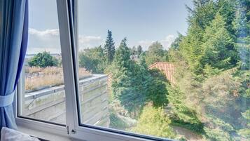 Quarto com uma janela grande com vista para uma área arborizada e um edifício com telhado vermelho.