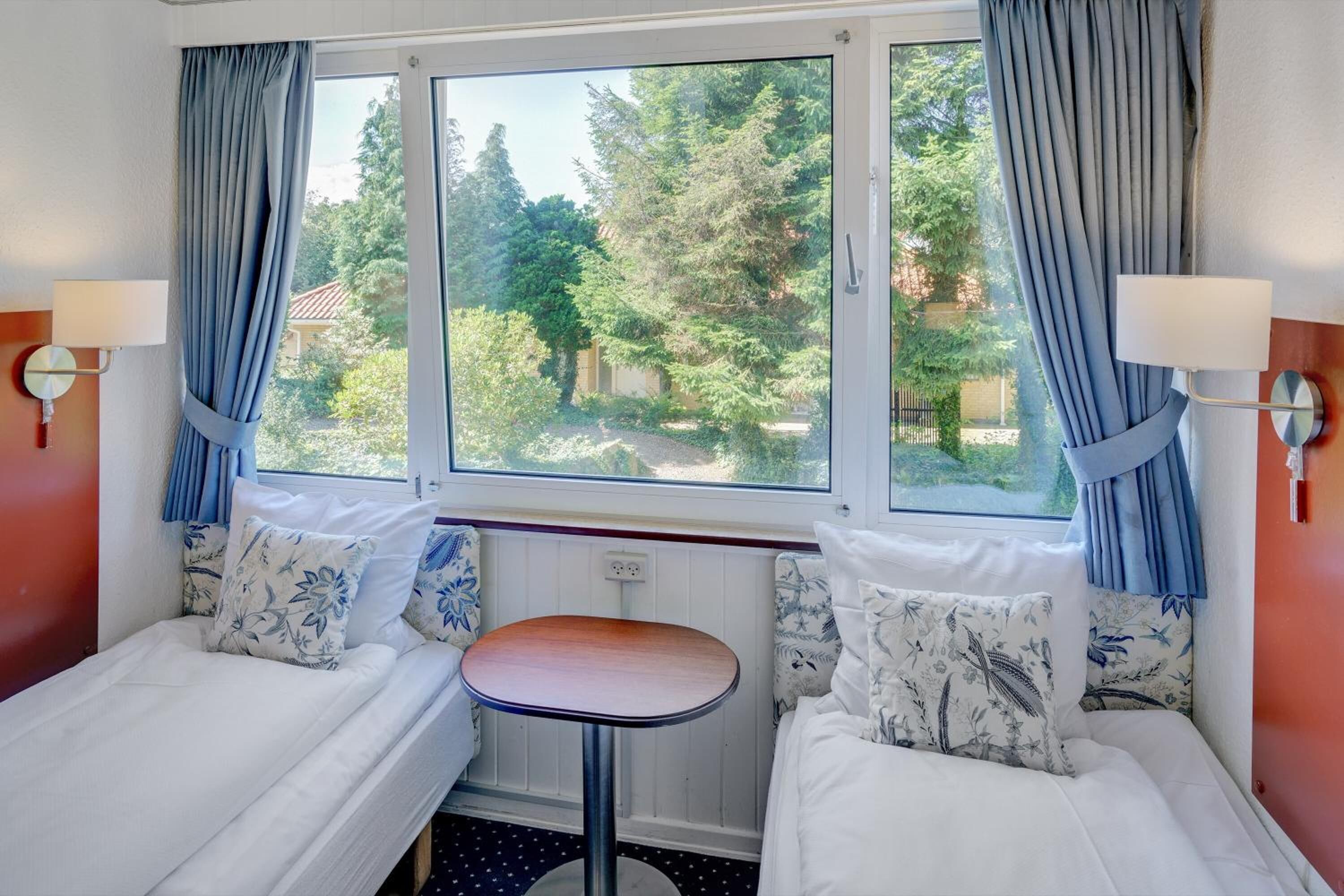 Ein Zimmer mit zwei Betten, einem kleinen Tisch und einem Fenster mit Vorhängen.