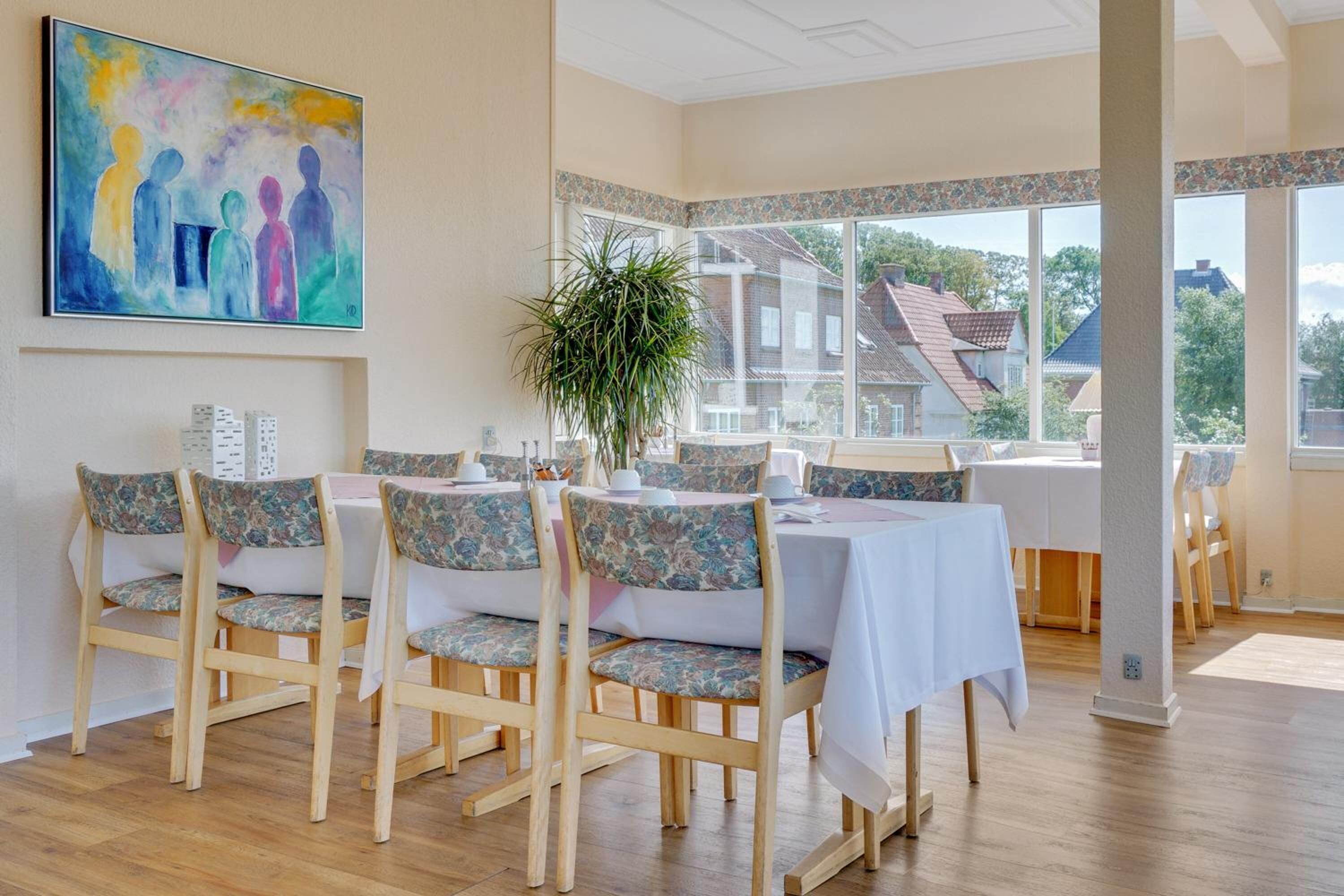A dining area with wooden chairs and tables, a large window with a view of houses, and a painting on the wall.
