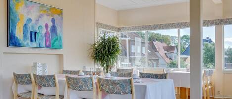 A dining area with wooden chairs and tables, a large window with a view of houses, and a painting on the wall.