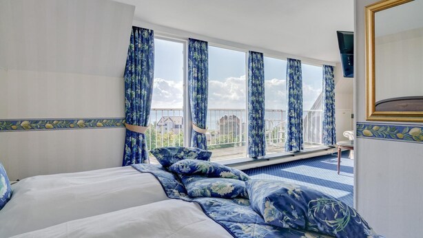 Habitación de hotel con una cama grande, vistas de la ciudad a través de ventanales del suelo al techo y un balcón con cortinas azules.