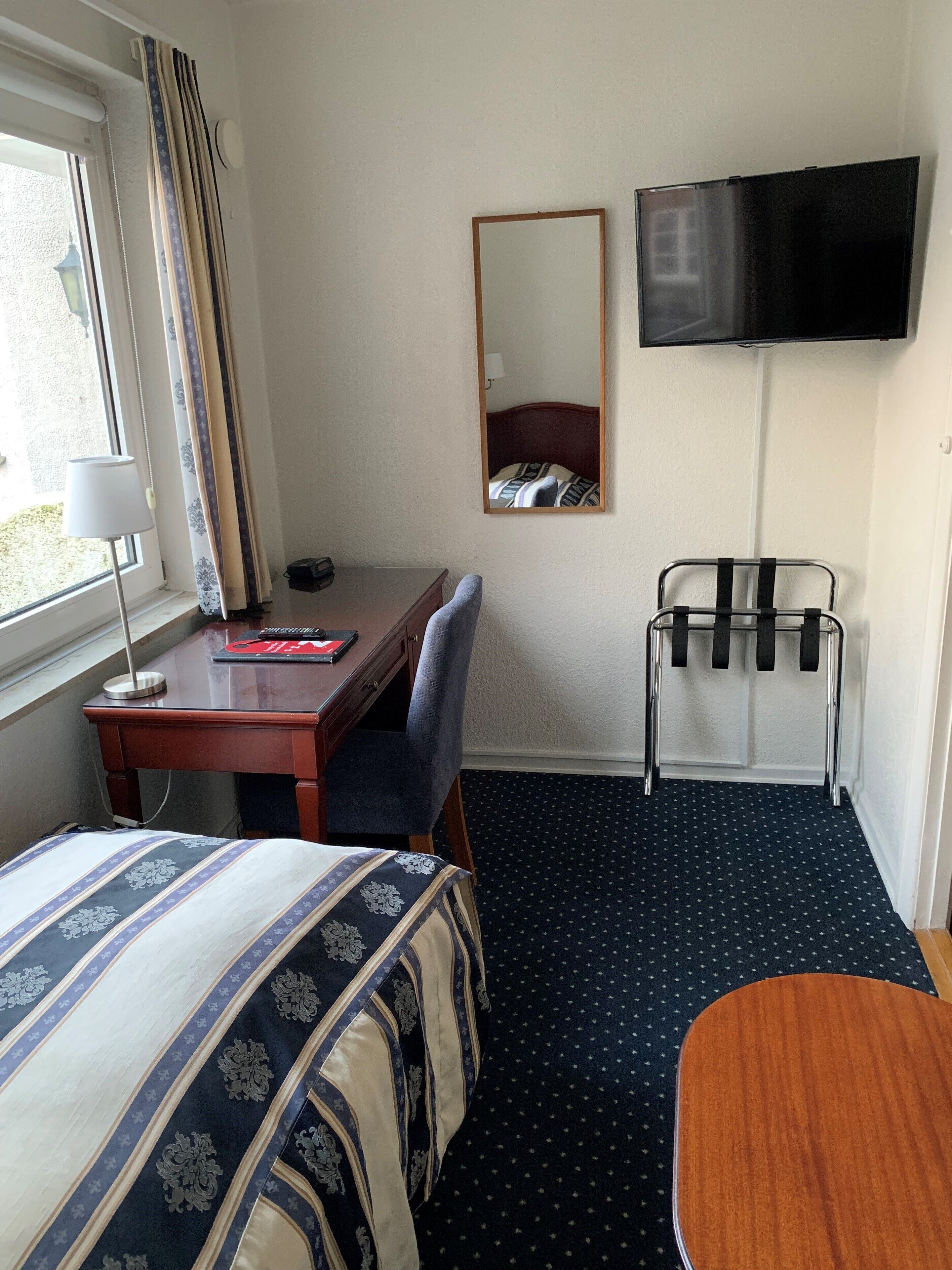 Habitación de hotel con cama, escritorio, silla, televisor montado en la pared y ventana con cortinas.