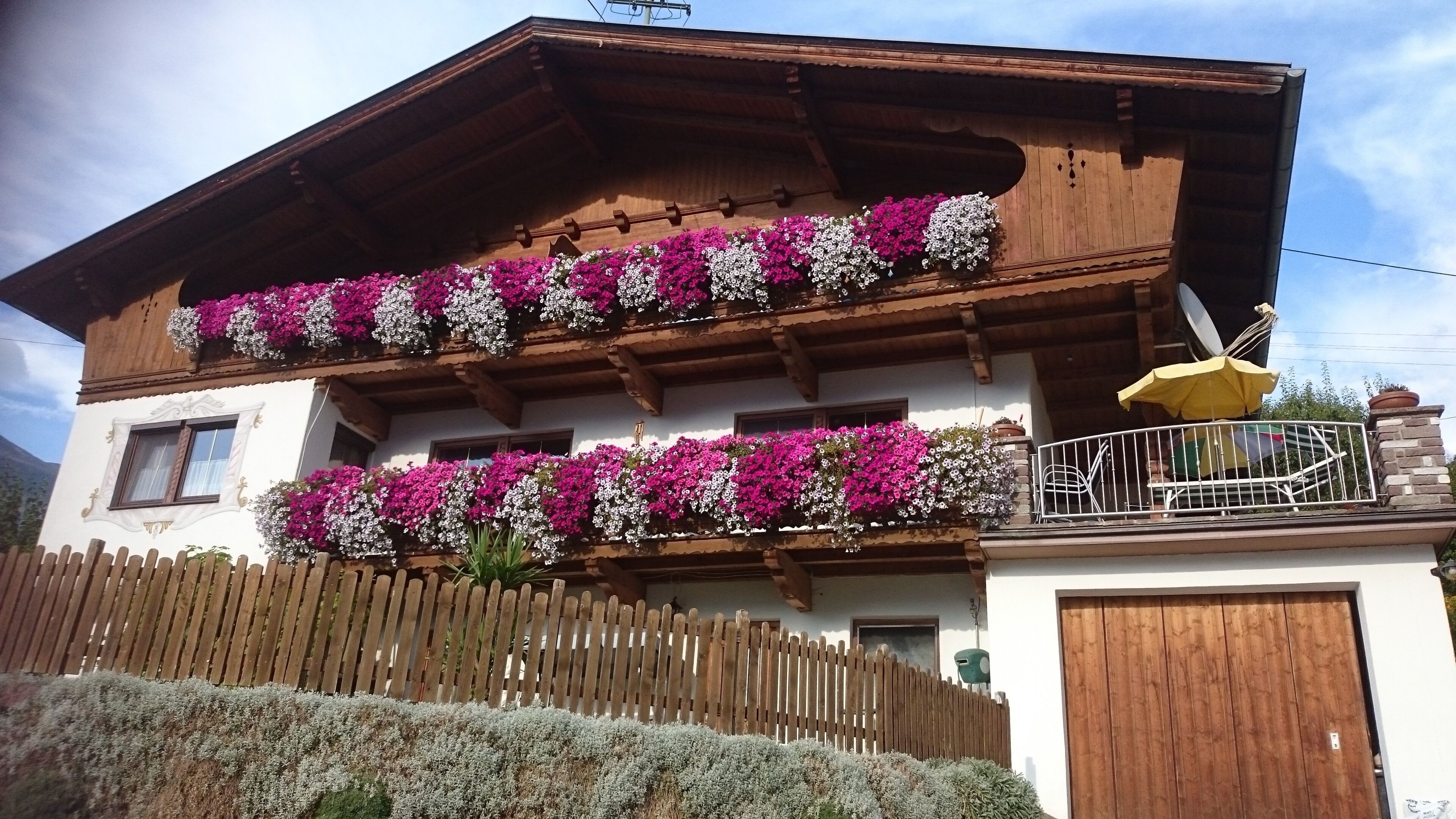 Ein traditionelles Chalet mit hölzernen Balkonen, die mit Blumen geschmückt sind, einem Holzzaun und einem Berg im Hintergrund.