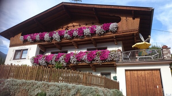 A traditional chalet with wooden balconies adorned with flowers, a wooden fence, and a mountain backdrop.