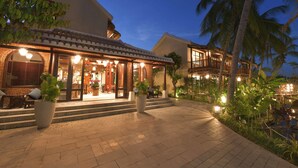 Una entrada de hotel bien iluminada con un patio cubierto, palmeras y una zona de piscina.
