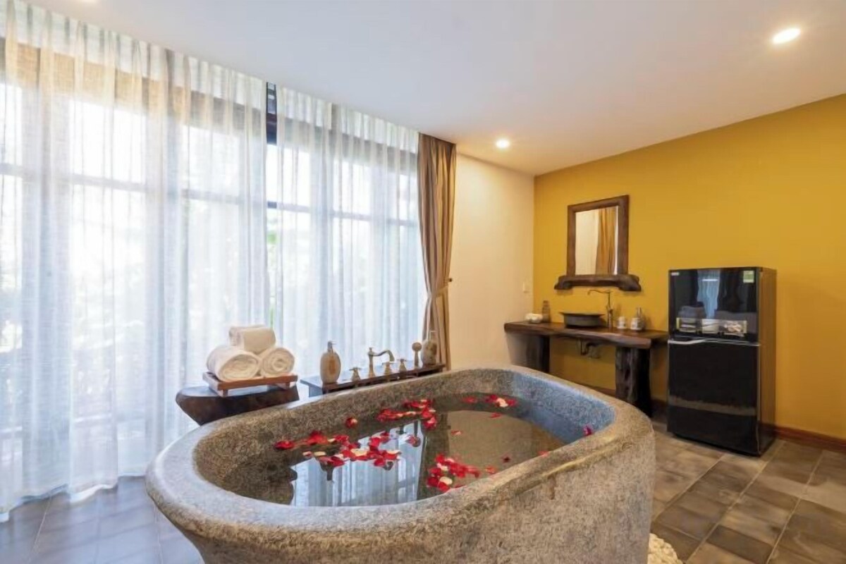 Una bañera de piedra con pétalos de rosa, un televisor y un refrigerador en una habitación con paredes amarillas y un ventanal grande.