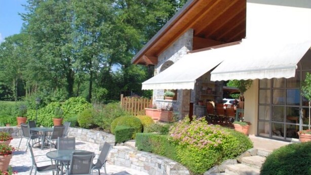 A stone building with a white exterior and wooden roof, featuring a patio with outdoor seating and potted plants.