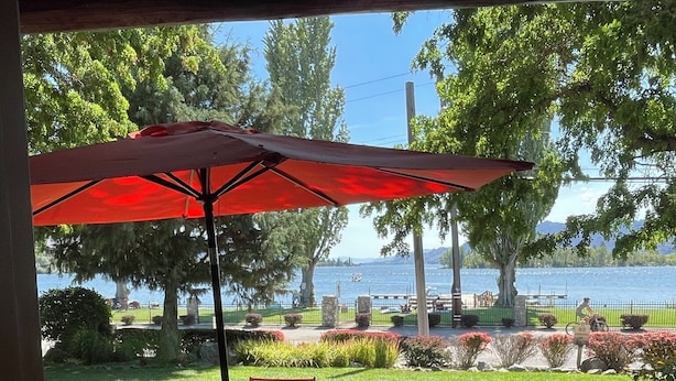 An outdoor patio with a glass table and red chairs, a red umbrella, and a view of a lake and trees. - Luxury Lakefront Townhouse 3Bed,3Bath Pool•Hot Tub•Beach - Canada's warmest lake (Osoyoos)