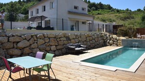 A pool area with a stone wall, a wooden deck, and a white building in the background.