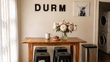 A kitchen with a wooden table, two black stools, a coffee cup, a plate with pastries, a vase with flowers, and a wall-mounted picture of a cow.