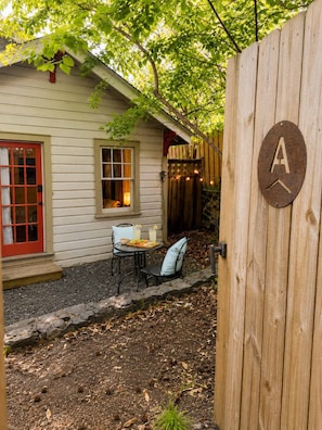 A wooden gate with a letter 'A' sign, a house with a red door, a window with a lit lamp, and a table with chairs outside.