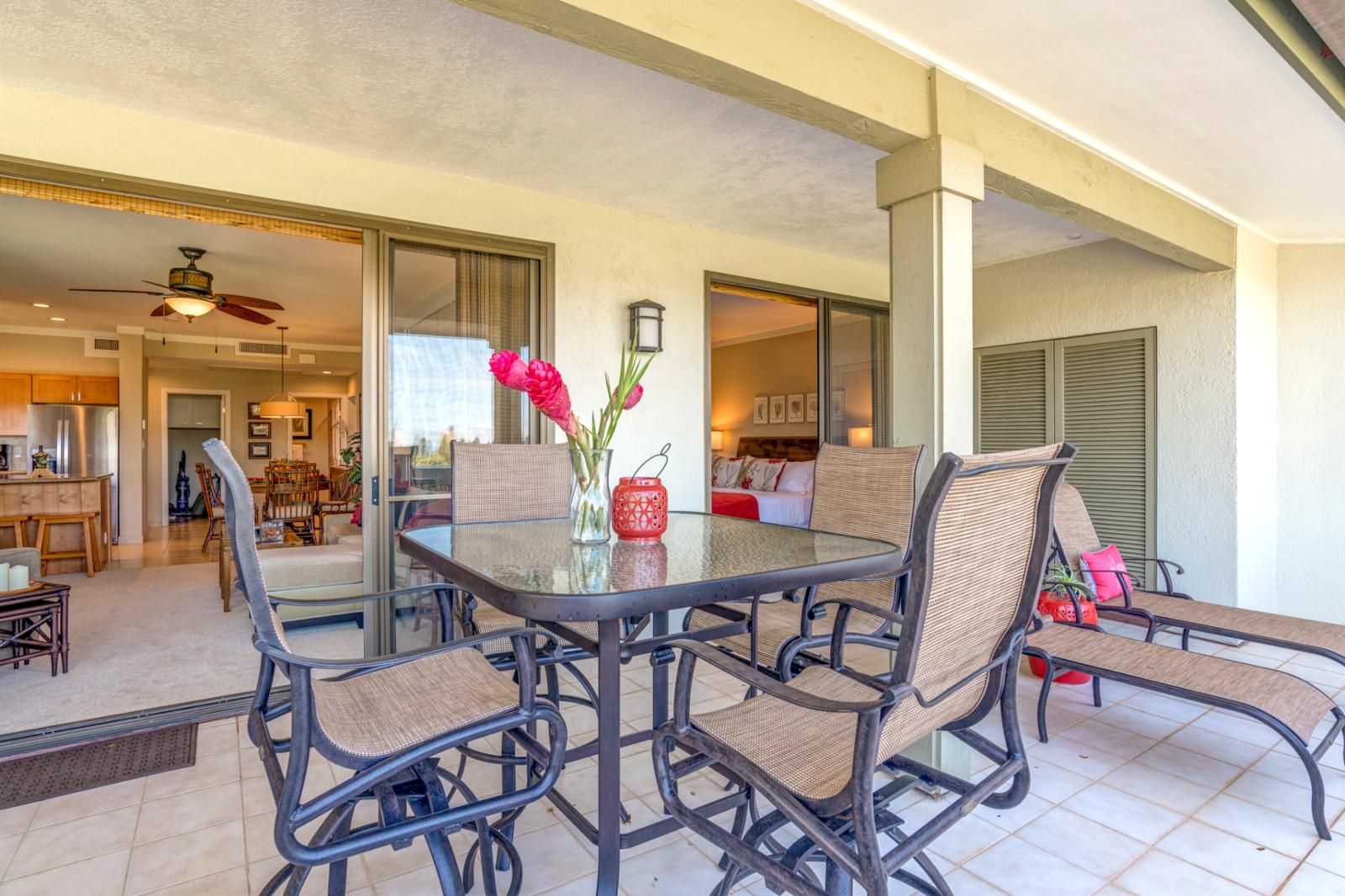 A dining area with a glass table and wicker chairs, a kitchen in the background, and a view of the living area through a glass door.