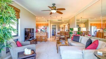 A spacious living room with a sofa, coffee table, and a television. There is a kitchen area with wooden cabinets and a dining table.