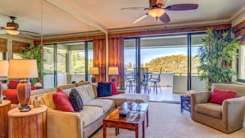 A spacious living room with a sofa, coffee table, and chairs, large windows, and a ceiling fan.