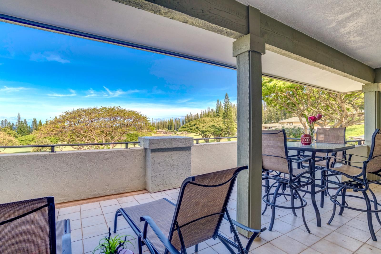 Eine überdachte Terrasse mit Gartenmöbeln, einem Tisch mit Blumenvase und Blick auf Golfplatz und Bäume.