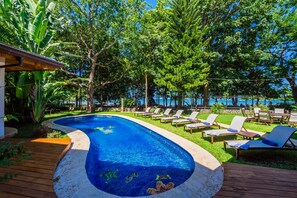 A swimming pool with a wooden deck, surrounded by lounge chairs and trees. - Beachfront 10BR Luxury Villa in Tamarindo – Walk to Town & Surf (Tamarindo)
