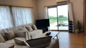 Ein modernes Wohnzimmer mit einer Couch, einem Couchtisch und einem Fernseher. Ein Schiebetüren-Glasfenster führt in einen Außenbereich mit Blick auf Grünflächen.