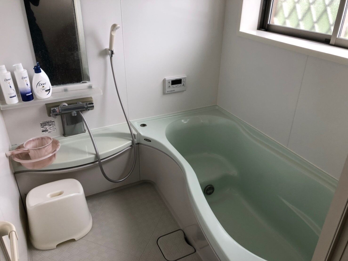 A bathroom with a green bathtub, a handheld showerhead, a sink with a soap dispenser, and a window with blinds.